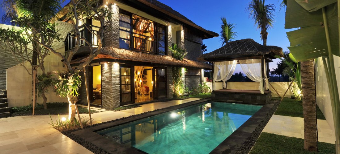 Modernes, zweistöckiges Ferienhaus im tropischen Stil mit Pool im Garten. Abendstimmung, warmes Licht aus dem Inneren, daneben eine offene Lounge mit weißen Vorhängen unter einem Pavillon.