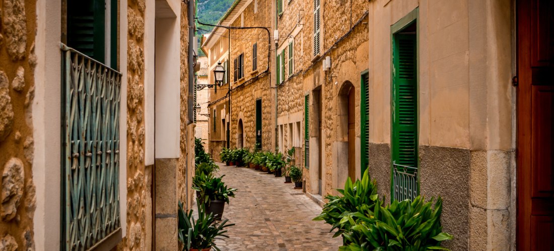 Eine malerische Altstadtstraße auf Mallorca, gesäumt von traditionellen Steinhäusern mit grünen Fensterläden und üppigen Topfpflanzen. Kopfsteinpflaster bedeckt den Boden, und im Hintergrund sind Berge zu sehen. Die Straße wirkt ruhig und einladend.