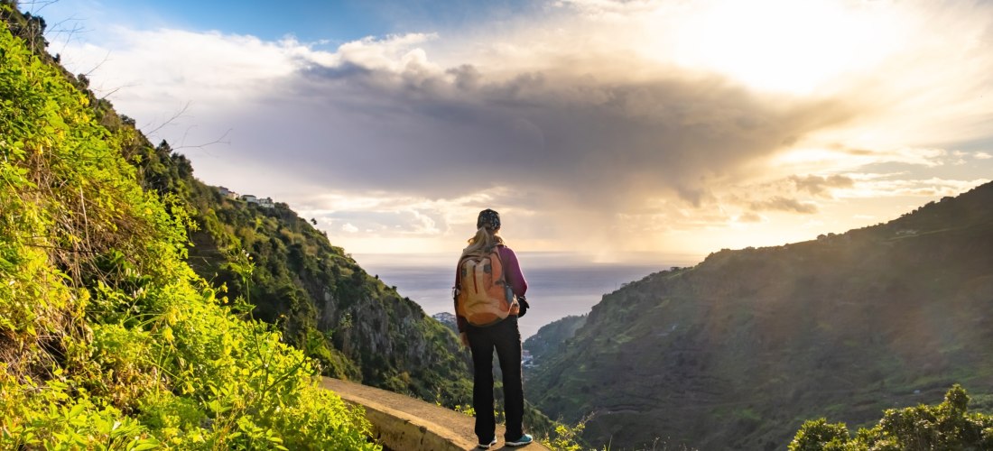Eine Frau mit Rucksack steht auf einem Levada-Weg auf Madeira und blickt über ein grünes Tal in Richtung Meer. Die Sonne bricht dramatisch durch die Wolken und beleuchtet die hügelige Landschaft mit warmem Licht.