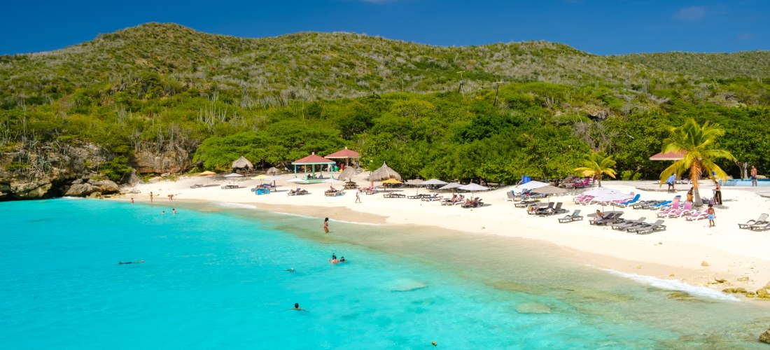 Breiter Sandstrand auf Curaçao mit zahlreichen Sonnenliegen und Sonnenschirmen vor türkisblauem Meer. Einige Menschen baden oder entspannen am Strand. Im Hintergrund grüne Hügel mit dichter Vegetation unter tiefblauem Himmel