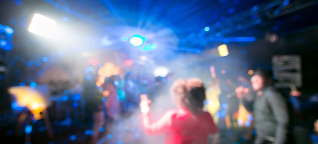 Verschwommene Aufnahme einer bunten Clubszene mit Tanzenden, Lichtstrahlen und Nebel – ausgelassene Partystimmung auf der Tanzfläche.