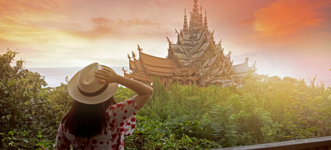 Frau mit Hut und Rucksack steht auf einem Balkon und blickt auf das kunstvoll geschnitzte Holzgebäude Sanctuary of Truth in Pattaya bei Sonnenuntergang. Im Vordergrund dichter grüner Wald, dahinter das Meer im Abendlicht