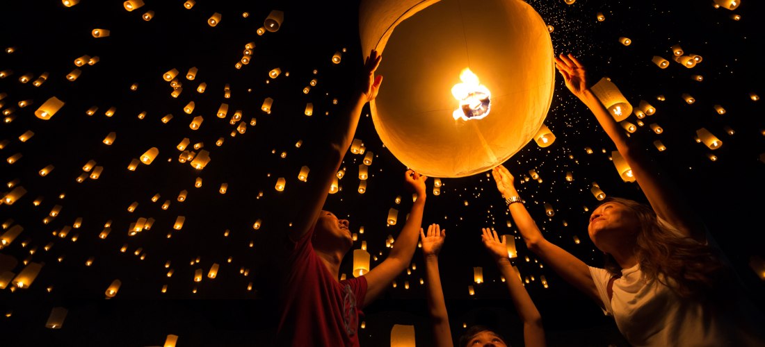 Das Bild zeigt eine bewegende Szene bei einem Himmelslaternen-Festival in Thailand: Eine Familie – bestehend aus einem Mann, einer Frau und einem Kind – lässt gemeinsam eine große, leuchtende Laterne in den Nachthimmel steigen. Um sie herum sind hunderte 