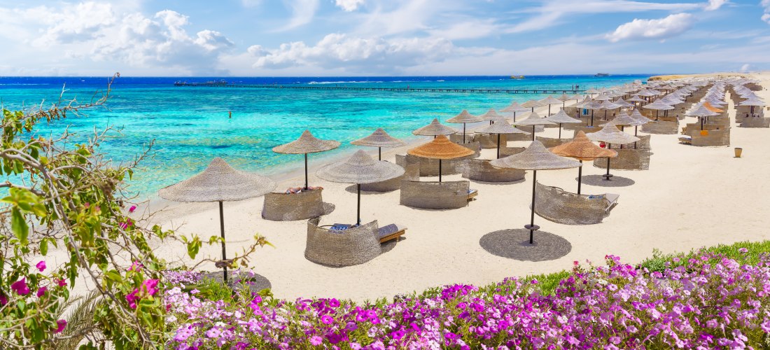 Breiter Sandstrand mit zahlreichen Strohliegen und Sonnenschirmen vor türkisblauem Meer. Im Vordergrund leuchtend pinke Blumen, im Hintergrund ein Holzsteg im Wasser. Der Himmel ist klar mit weißen Wolken.