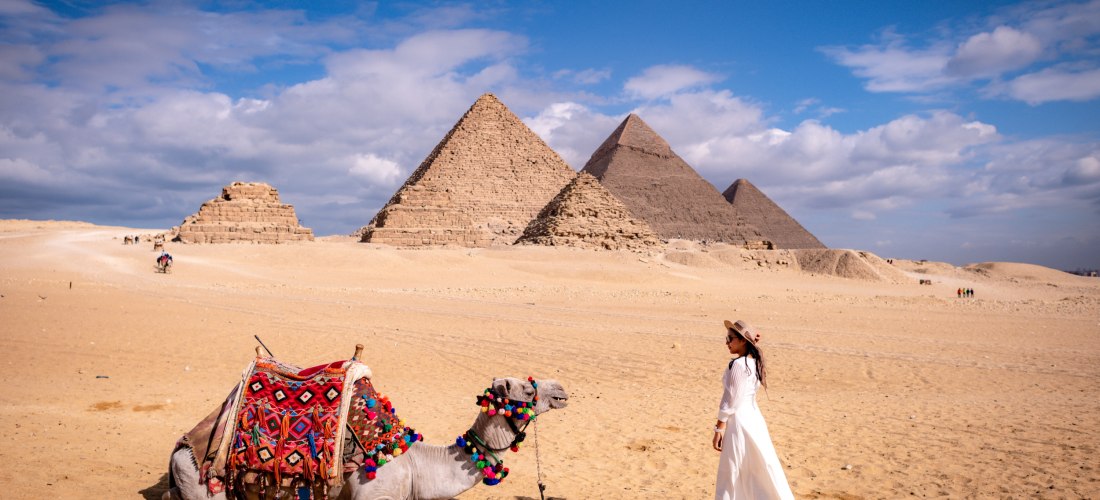Das Bild zeigt eine Frau in einem weißen Kleid mit Sonnenhut, die einem geschmückten Kamel gegenübersteht. Das Kamel liegt im Wüstensand und trägt eine bunte Decke mit traditionellem Muster. Im Hintergrund erheben sich die bekannten Pyramiden von Gizeh un