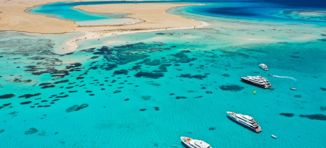 Drohnenaufnahme von mehreren luxuriösen Yachten, die in einer türkisblauen Lagune vor einer Sandinsel im Roten Meer ankern. Im Wasser sind dunkle Korallenriffe zu sehen, im Hintergrund weite Sandflächen und das tiefblaue offene Meer.