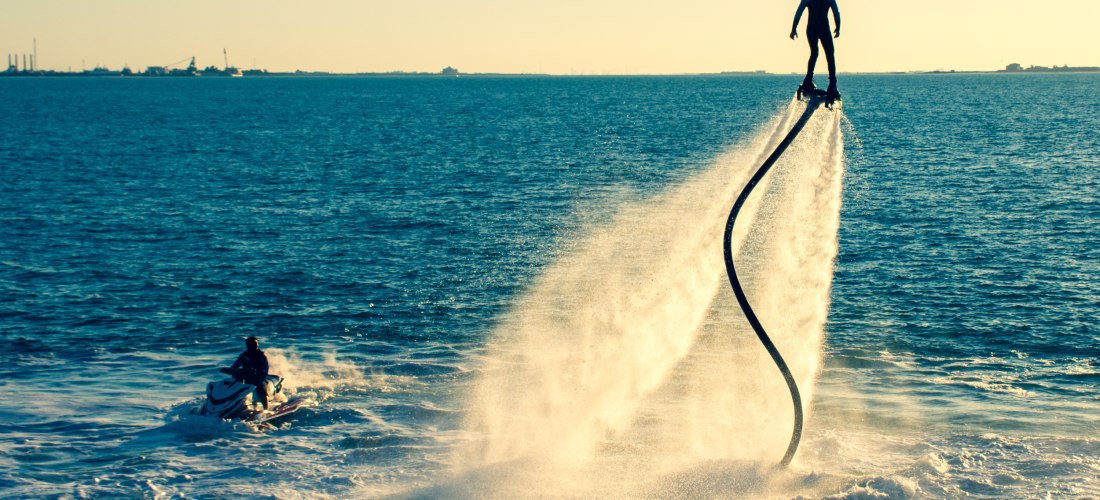 Person schwebt auf einem Flyboard über dem Meer, angetrieben von Wasserstrahlen, mit Jet-Ski im Hintergrund