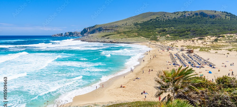 Weitläufiger Sandstrand Cala Mesquida auf Mallorca mit hohen Wellen, Liegen und Sonnenschirmen sowie grünen Hügeln im Hintergrund
