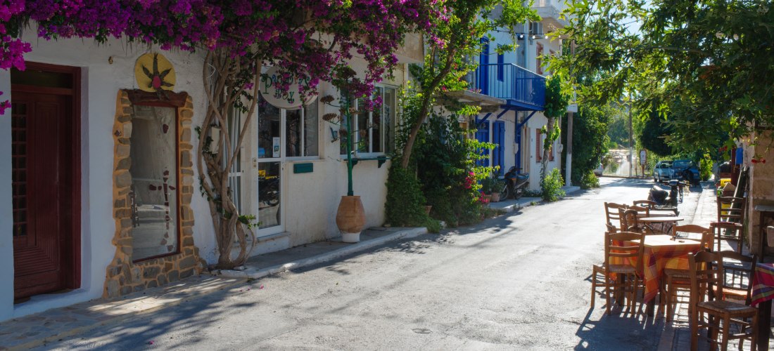 Schmale mediterrane Dorfstraße mit weißen Häusern, üppig blühender Bougainvillea, kleinen Geschäften und einem Straßencafé mit Holztischen und Stühlen im Schatten