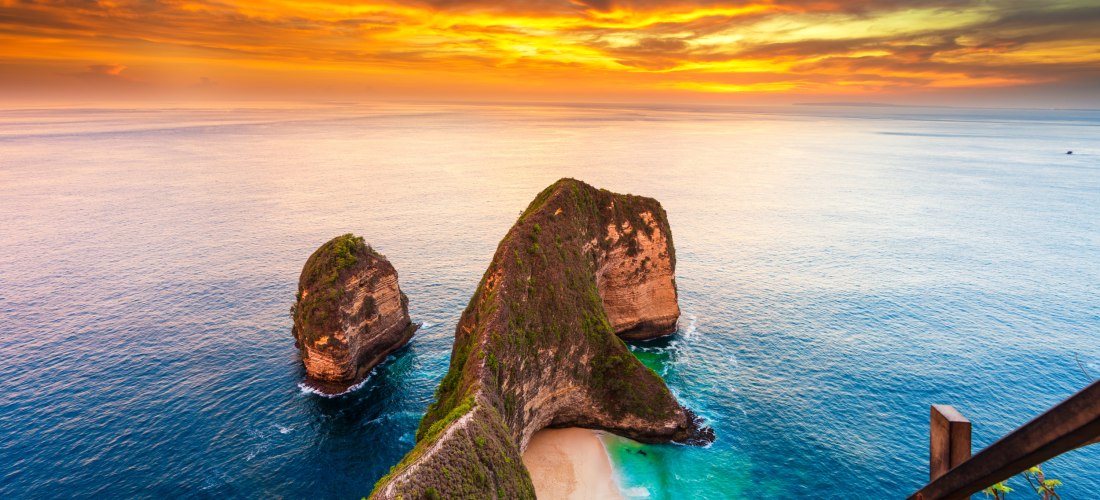 Spektakuläre Aussicht auf den berühmten Kelingking Beach auf der indonesischen Insel Nusa Penida bei Sonnenuntergang. Im Vordergrund führt ein schmaler Holzpfad mit Geländer hinab zur sichelförmigen Bucht mit weißem Sand und türkisfarbenem Wasser. Die mar