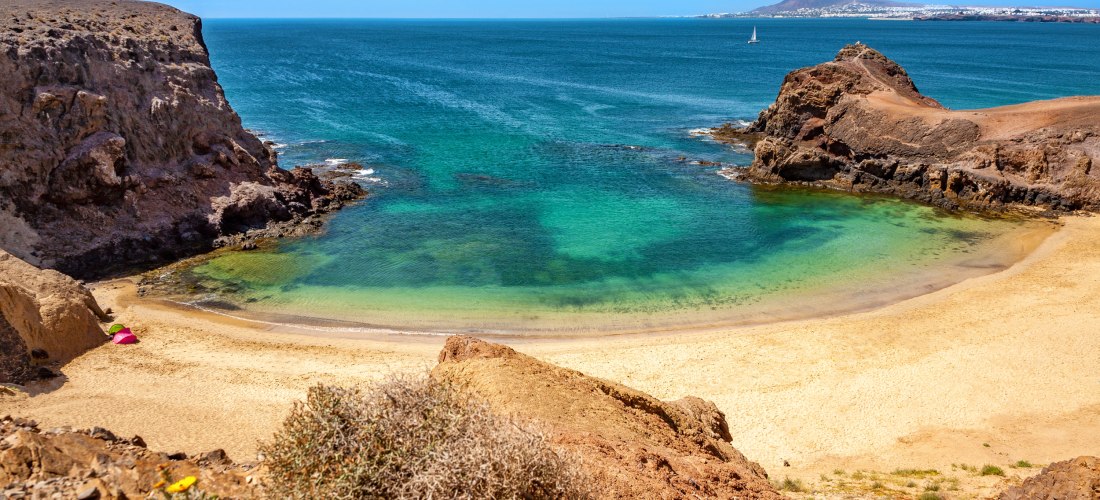Kleine, abgelegene Sandbucht mit türkisfarbenem Wasser, umgeben von Felsen. Am Strand liegen zwei bunte Sonnenschirme, im Hintergrund ist das offene Meer zu sehen.