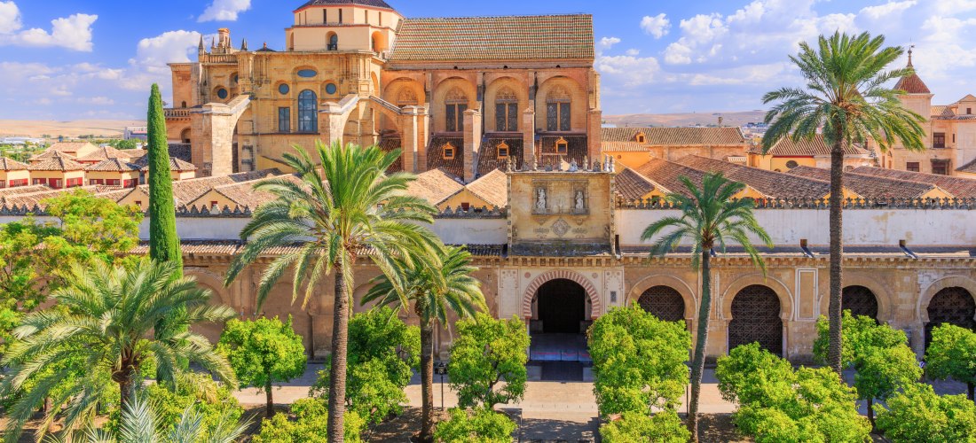 Blick auf die Mezquita-Kathedrale von Córdoba mit maurisch-christlicher Architektur. Im Vordergrund ein grüner Orangenhof mit Palmen, im Hintergrund das imposante Gebäude mit Bögen, Zinnen und Türmen unter blauem Himmel mit weißen Wolken.