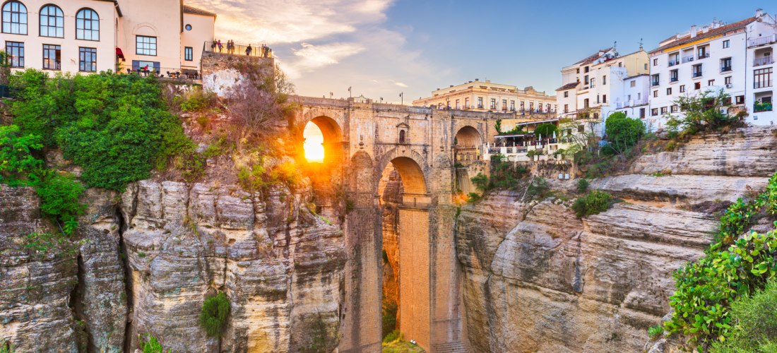 Die Puente Nuevo in Ronda bei Sonnenuntergang. Die beeindruckende Steinbrücke überspannt eine tiefe Schlucht und verbindet die Altstadt mit dem neueren Stadtteil. Sonnenlicht scheint durch den mittleren Brückenbogen.