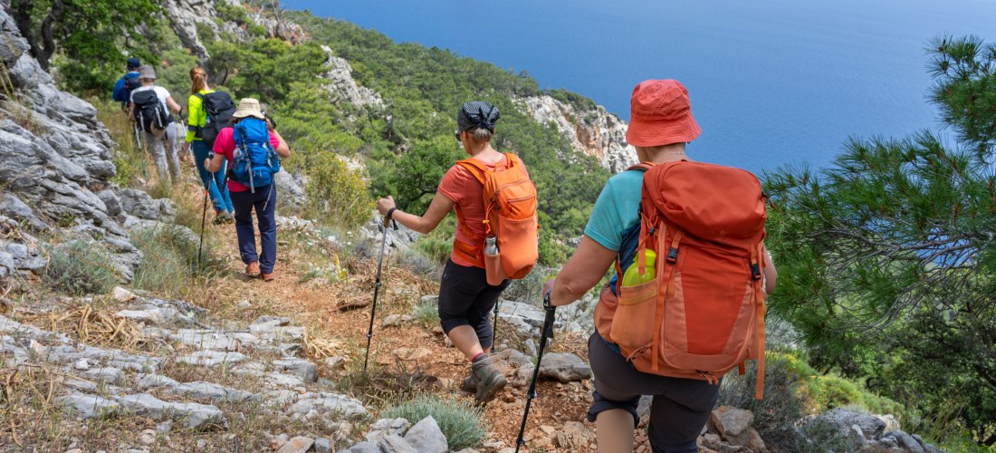 Entlang schmaler Pfade und steiler Felsen führt diese Wanderung durch die atemberaubende Küstenlandschaft der Türkischen Riviera. Eine Gruppe Wandernder genießt den Weitblick über das azurblaue Mittelmeer, mit einer kleinen Insel am Horizont. Natur, Beweg