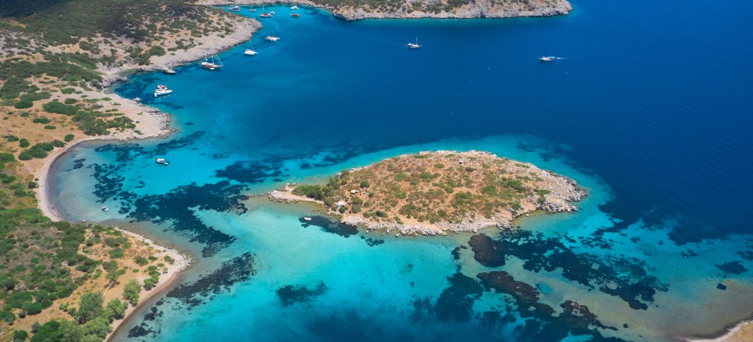 Diese atemberaubende Luftaufnahme zeigt eine idyllische Küstenlandschaft mit kristallklarem Wasser, kleinen Ankerbuchten und einer grünen Insel mitten im türkisblauen Meer. Zahlreiche Yachten und Segelboote liegen ruhig vor Anker – ein perfekter Ort zum E