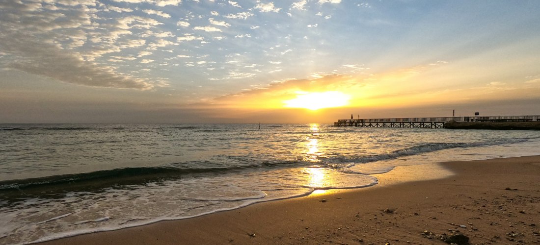 Sonnenuntergang am Meer mit ruhigen Wellen, die auf den Sand treffen. Rechts ist ein Holzsteg, der ins Wasser führt. Der Himmel ist mit gold-blauen Wolken durchzogen.