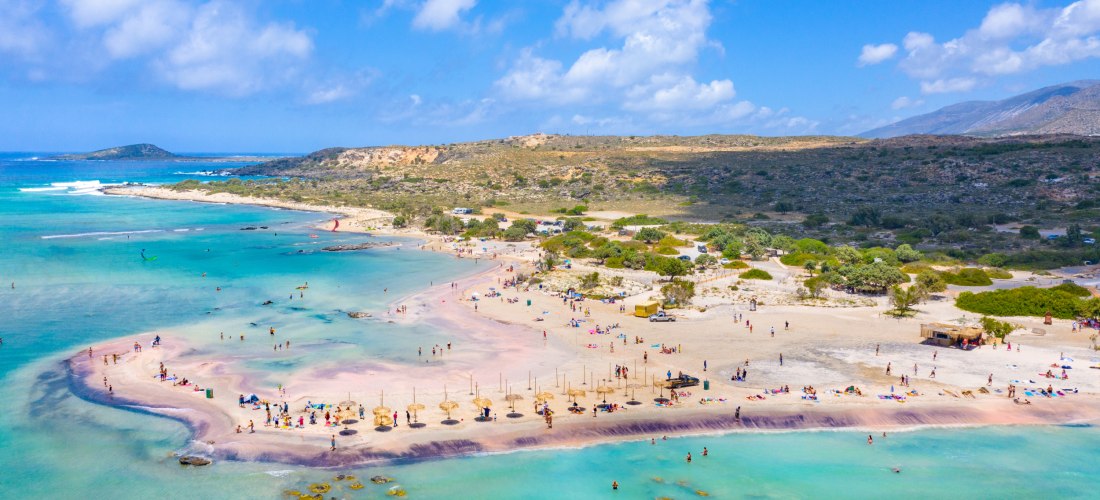 Der berühmte Strand von Elafonissi auf Kreta mit rosa schimmerndem Sand, türkisfarbenem Wasser und flachen Lagunen. Viele Menschen genießen den Tag am Strand, einige baden, andere entspannen unter Strohdächern. Die Landschaft im Hintergrund ist leicht hüg