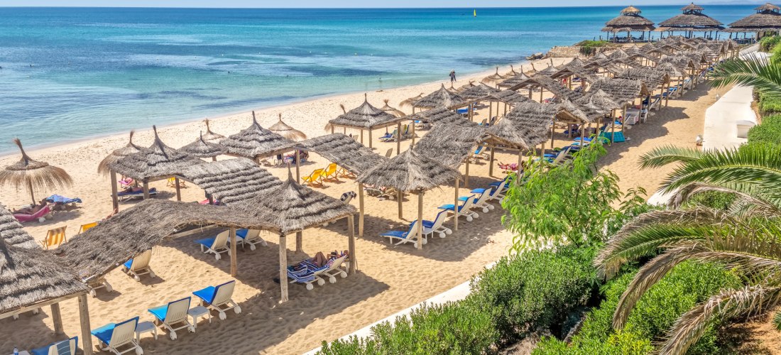 Ein sonniger Sandstrand mit zahlreichen Sonnenliegen unter strohgedeckten Sonnenschirmen. Im Hintergrund erstreckt sich das türkisfarbene Meer bis zum Horizont. Einige Menschen entspannen im Schatten, andere baden im ruhigen Wasser. Im Vordergrund sind gr