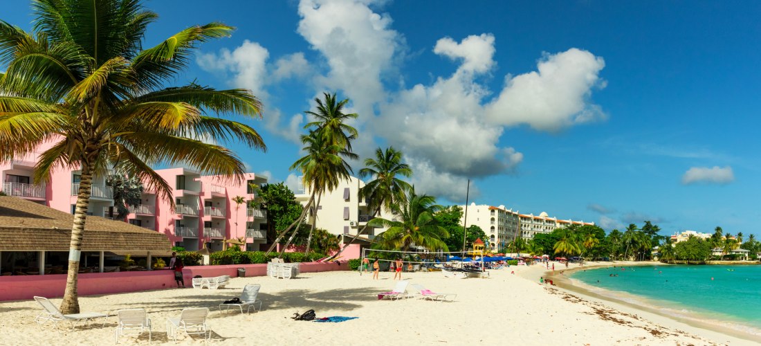 Blick auf einen hellen Sandstrand mit Palmen, Sonnenliegen und mehreren Hotels direkt am Meer. Im Vordergrund stehen rosa und weiße Hotelgebäude, dahinter Volleyballnetze und bunte Sonnenschirme. Das Meer ist türkisblau und der Himmel zeigt ein paar weiße