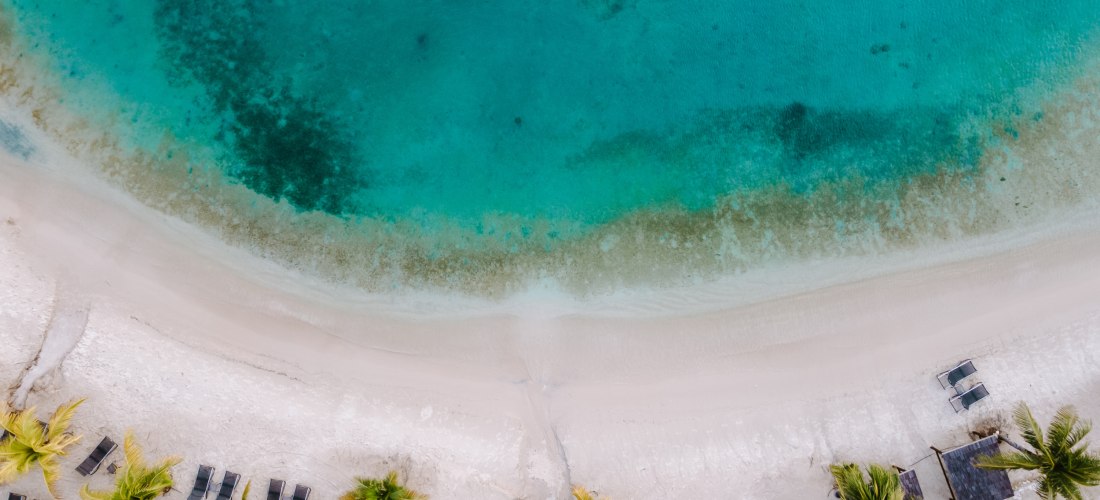 Drohnenaufnahme eines tropischen Strandes mit weißem Sand, türkisblauem Meer und Palmen. Am unteren Bildrand sind Liegen und kleine strohgedeckte Hütten zu sehen, das Wasser zeigt verschiedene Blautöne je nach Tiefe und Untergrund.