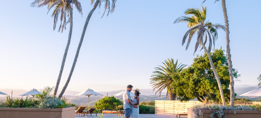 Ein Paar steht in romantischer Pose an einem kleinen Teich, eingerahmt von Palmen, mit Blick auf ein luxuriöses Resort im Hintergrund. Die Szene spielt sich bei klarem Himmel und weichem Abendlicht ab – perfekte Urlaubsstimmung.
