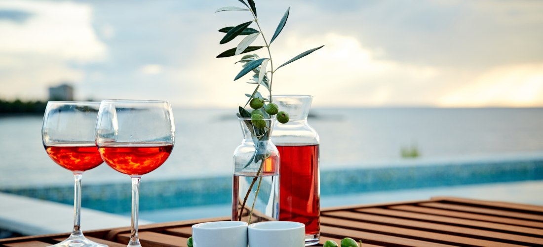 Zwei Gläser Roséwein, Oliven und eine Karaffe auf einem Holztisch mit Meerblick bei Sonnenuntergang