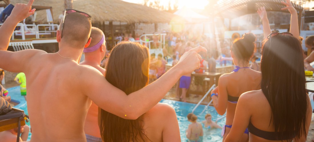Menschen feiern eine lebhafte Poolparty bei Sonnenuntergang. Ein junger Mann und eine Frau in Badekleidung stehen Arm in Arm am Poolrand, während viele andere im Wasser und auf der Terrasse tanzen und feiern. Im Hintergrund sind Palmen, Strohdächer und ei