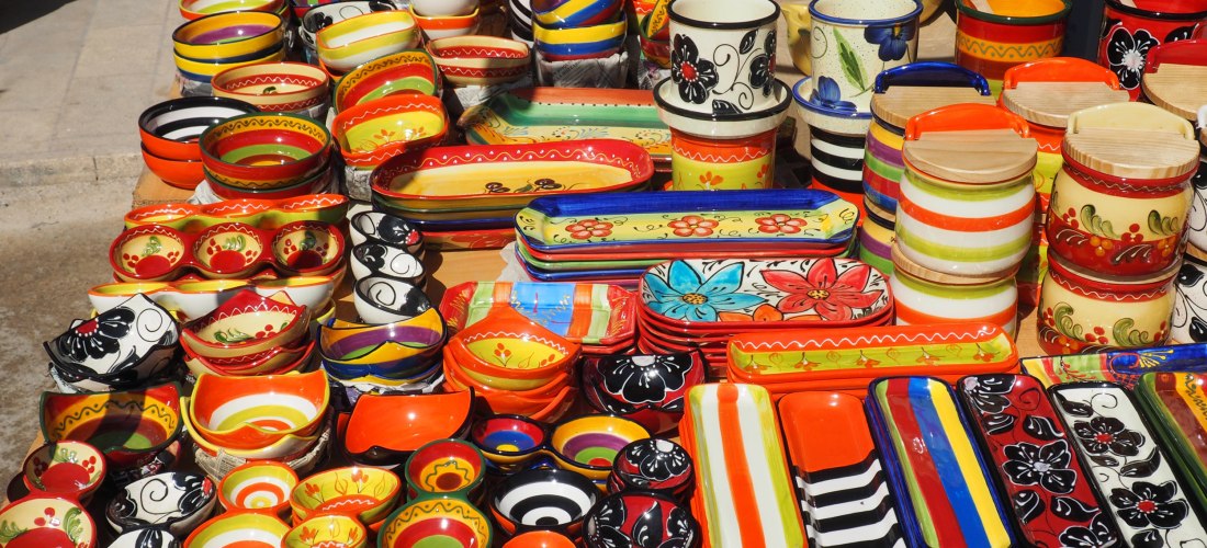 Keramikstand auf einem Markt mit einer Vielzahl an bunt bemalten Schalen, Tellern und Bechern. Die Muster sind farbenfroh mit Blumen, Streifen und Ornamenten in kräftigem Rot, Gelb, Orange und Schwarz