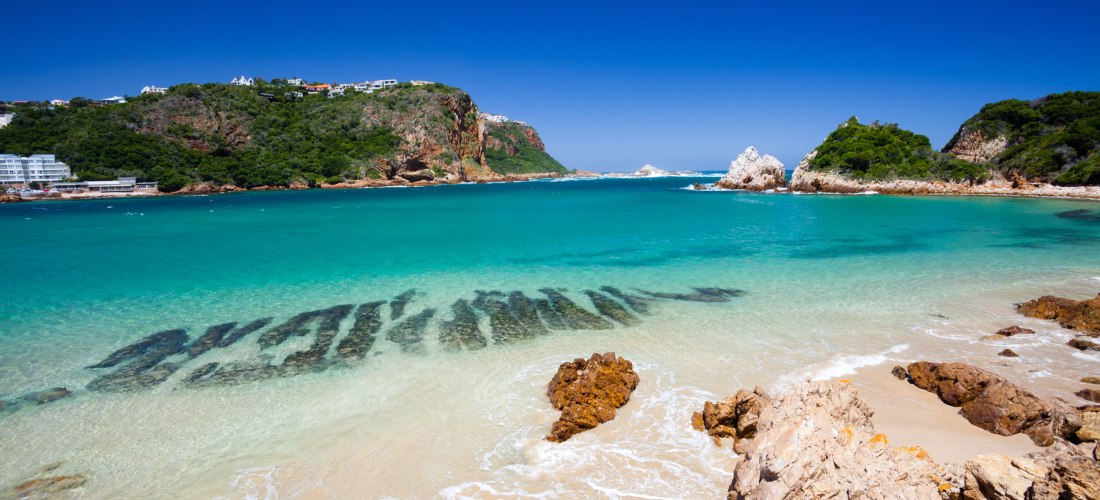 Blick auf einen traumhaften Strand in Südafrika mit türkisblauem Wasser und feinem Sand. Im Hintergrund erheben sich grüne Klippen, die teilweise von weißen Gebäuden gesäumt sind. Der Himmel ist strahlend blau – perfektes Strandwetter.