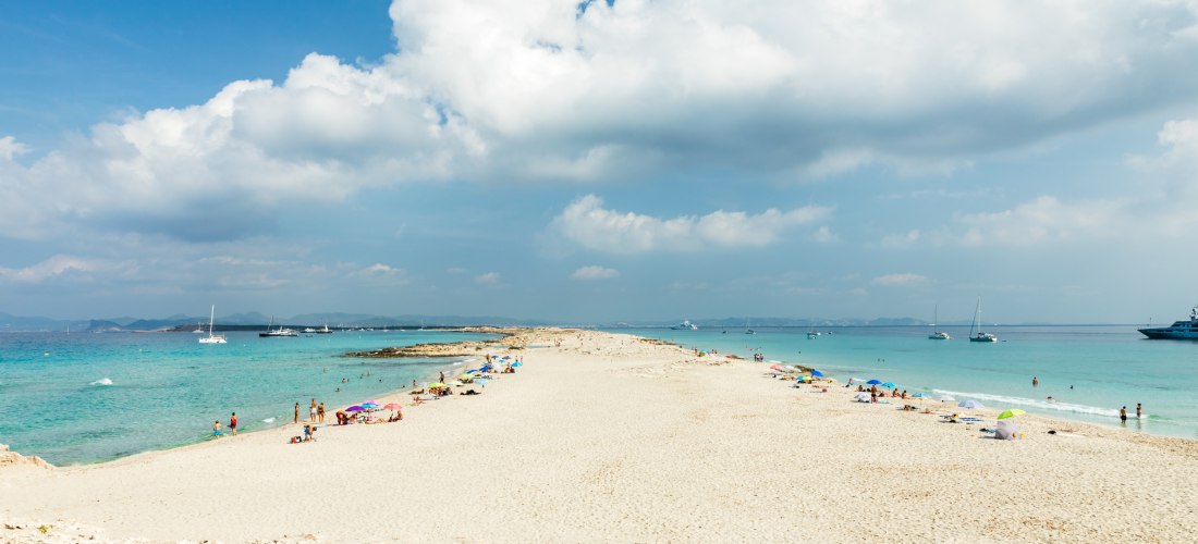 Blick auf die Playa de Ses Illetes auf Formentera mit weißem Sandstrand, Dünen und türkisgrünem Meer. Mehrere Yachten und Boote liegen vor der Küste, im Hintergrund sind Berge sichtbar.