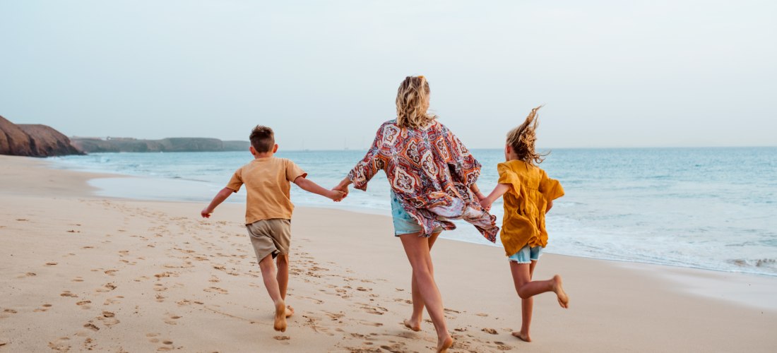 Frau läuft barfuß mit zwei Kindern an der Hand am Strand entlang. Der Sand ist weich, das Meer ruhig und der Himmel hell
