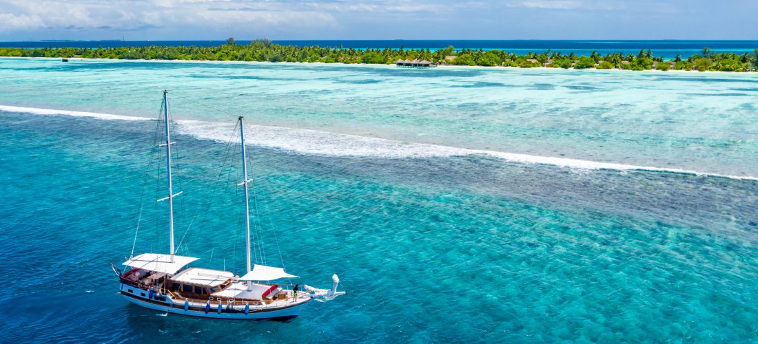 Ein elegantes Segelboot gleitet durch das türkisfarbene Wasser einer tropischen Insel. Die sanften Wellen und die üppige Vegetation im Hintergrund schaffen eine traumhafte Kulisse für ein unvergessliches Segelerlebnis.