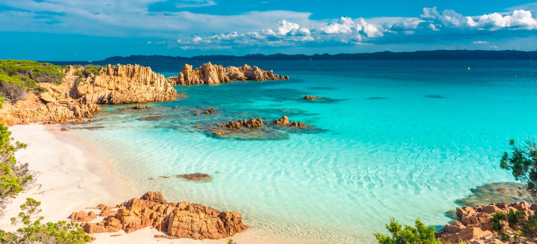 Idyllische Bucht auf Sardinien mit weißem Sandstrand, türkisfarbenem Wasser und markanten orangefarbenen Felsen. Im Vordergrund rahmen grüne Sträucher die Szene ein, während im Hintergrund das tiefblaue Meer und eine ferne Küstenlinie zu sehen sind.