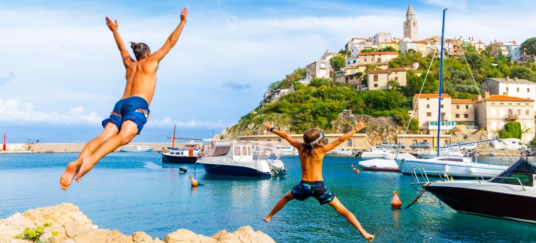 Ein Vater und sein Kind springen gleichzeitig von einem Felsen ins Meer. Im Hintergrund liegt ein kleiner Küstenort mit bunten Häusern am Hang, Booten im Wasser und einem Hafen.