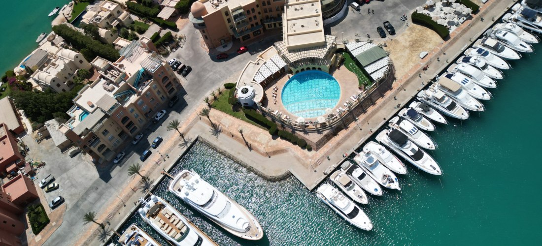 Luftaufnahme einer Marina mit einem großen runden Pool, umgeben von modernen Gebäuden, Palmen und einer Reihe weißer Yachten im türkisfarbenen Wasser.