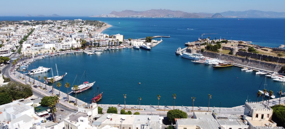 Panorama von Kos-Stadt: halbkreisförmiger Hafen mit Promenade, Yachten und Festungsanlage am rechten Ufer, dahinter die Ägäis