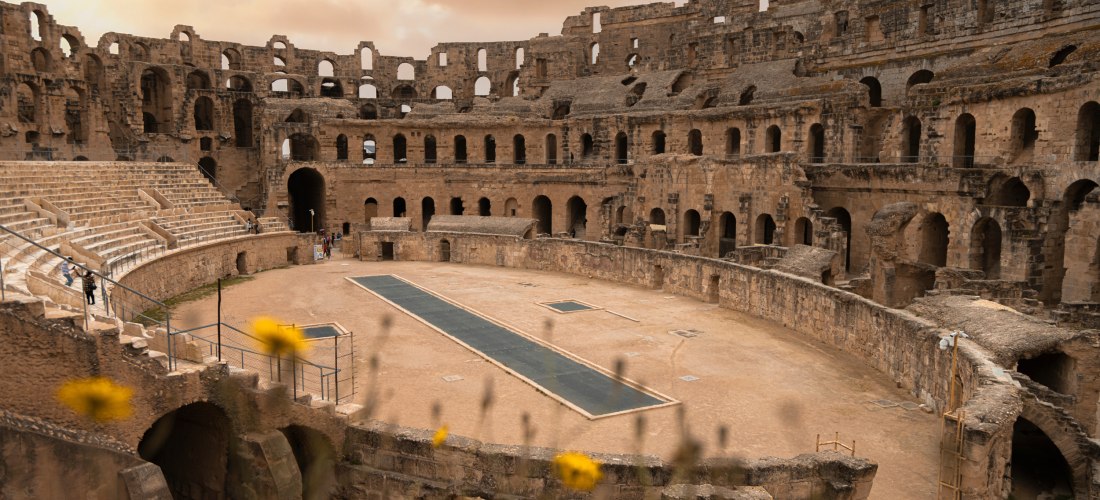 Blick ins römische Amphitheater von El Djem in Tunesien bei Sonnenuntergang. Die monumentalen Steinmauern des Kolosseums ragen in den Himmel, während im Vordergrund gelbe Blumen unscharf die Szene einrahmen. Einige Menschen schlendern durch das untere Run
