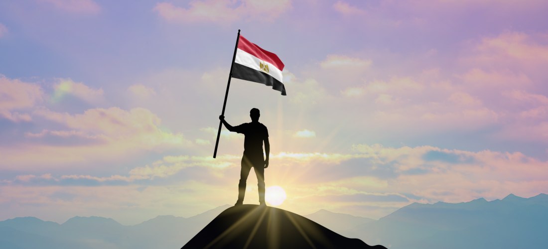 Eine Silhouette eines Mannes steht auf einem Berggipfel und hält die ägyptische Flagge. Die Sonne geht im Hintergrund auf, der Himmel ist rosa-blau gefärbt.