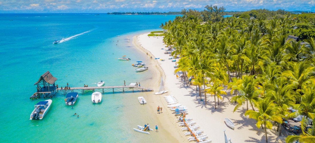Dieses atemberaubende Bild zeigt einen tropischen Traumstrand mit weichem, weißen Sand und einer üppigen Palmenlandschaft. Am Ufer reihen sich Wassersportgeräte wie Kajaks und Segelboote aneinander, während Menschen das klare, warme Wasser genießen. Ein l