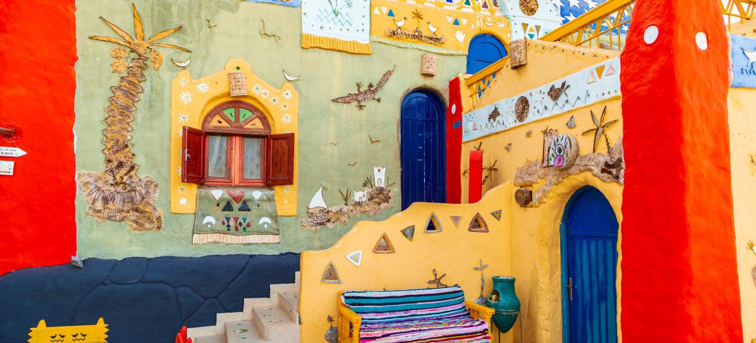 Farbenfrohe Fassade eines nubischen Hauses in Ägypten mit leuchtend roten, gelben und blauen Wänden. Die Wand ist reich mit traditionellen Mustern, Tieren, Symbolen und bemalten Fenstern verziert. Eine bunt gestreifte Holzbank steht im Vordergrund