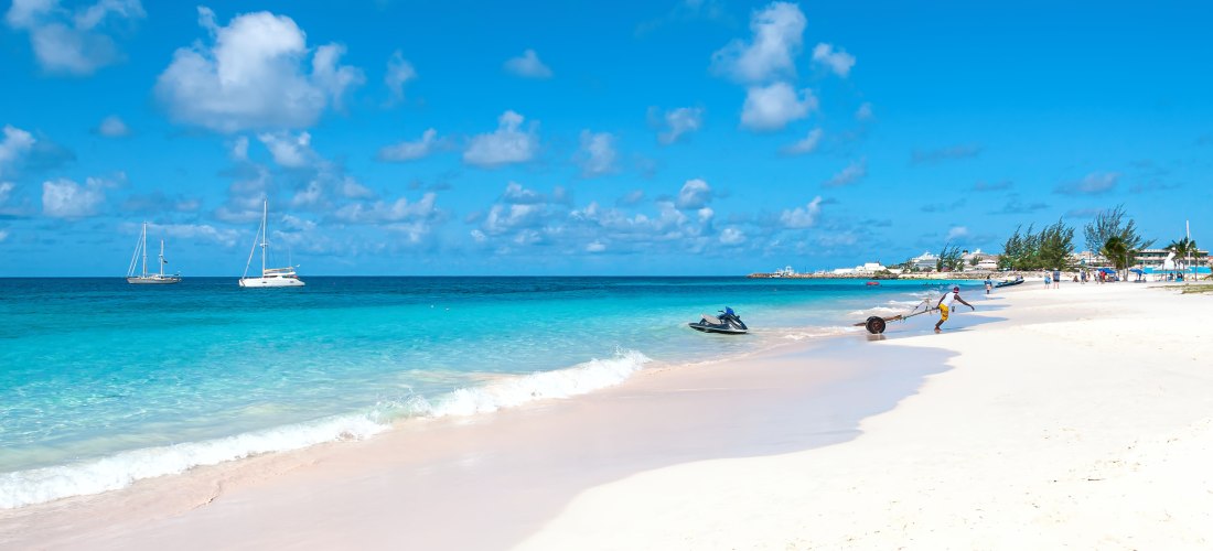 Weißer Sandstrand auf Barbados mit türkisblauem Meer, Jet-Ski und Segelbooten bei blauem Himmel.