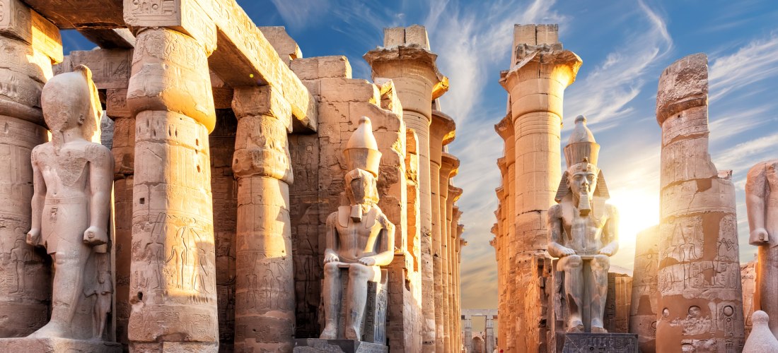 Blick auf den Luxor-Tempel in Ägypten mit großen Säulen und monumentalen Pharaonen-Statuen. Die Sonne geht hinter dem Tempel unter und taucht das Bauwerk in warmes Licht.