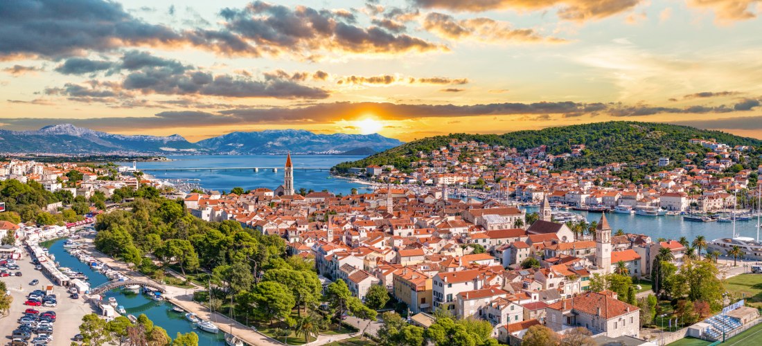 Luftaufnahme der Altstadt von Trogir in Kroatien. Die mittelalterliche Stadt liegt auf einer kleinen Insel, umgeben von türkisblauem Wasser. Zu sehen sind die historischen Gebäude mit roten Ziegeldächern, der markante Glockenturm der Kathedrale von Trogir