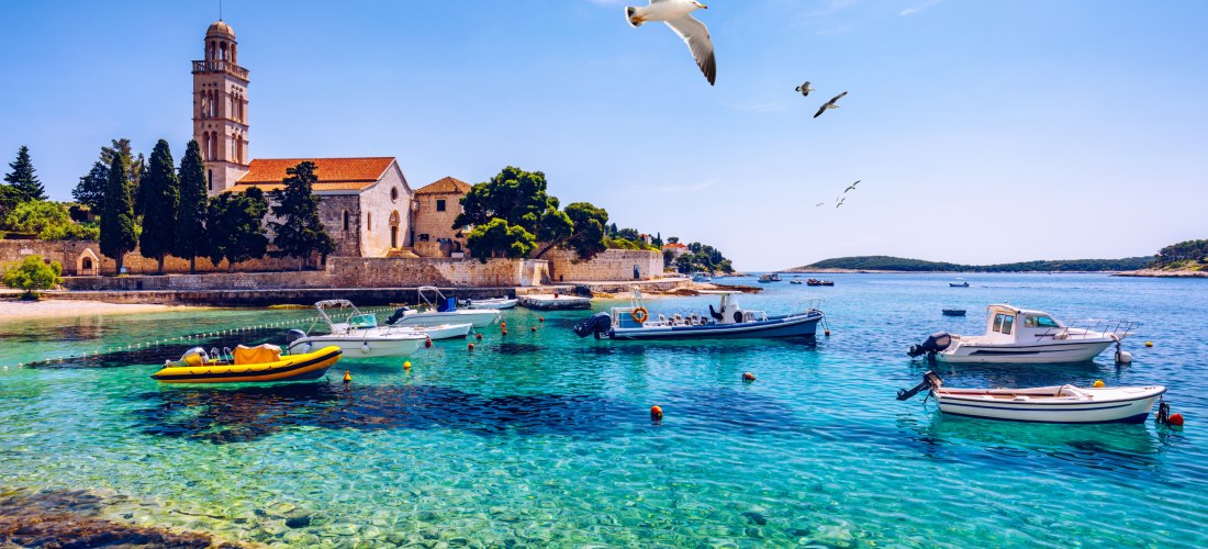 Hafenansicht mit klarem türkisfarbenem Wasser, kleinen Booten und einem alten Kloster mit rotem Dach und Glockenturm im Hintergrund. Möwen fliegen über das Wasser