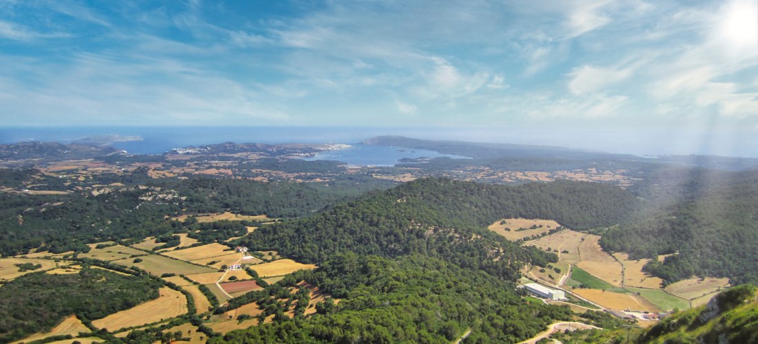 Weitläufiger Panoramablick über die hügelige Landschaft von Menorca. In der Ferne sind Felder, Wälder und die Küstenlinie mit dem Meer zu sehen. Die Szenerie ist von leicht bewölktem, sonnigem Himmel überzogen, mit weichem Licht von der rechten Seite.