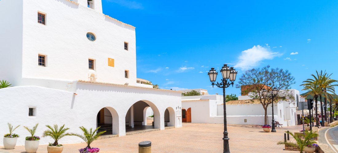 Weiße Kirche mit Arkaden und Glockenturm im Ort Sant Josep de sa Talaia auf Ibiza. Vor dem Gebäude befindet sich ein gepflasterter Platz mit Blumenkübeln, Laternen und Palmen unter blauem Himmel.
