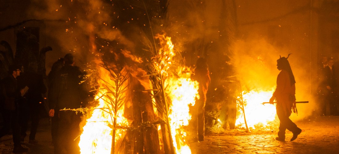 Nachtaufnahme eines traditionellen Festes mit mehreren brennenden Holzstapeln. Eine Person in einem roten Kostüm mit Hörnern, die einen Teufel darstellen, steht neben den Flammen. Weitere Menschen in dunkler Kleidung beobachten das Geschehen im Hintergrun