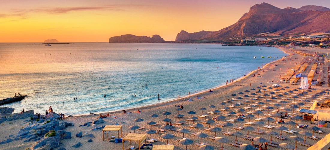 Ein langer Sandstrand auf Kreta bei Sonnenuntergang mit zahlreichen Sonnenliegen und Schirmen. Die Küste ist von sanften Wellen umspült, während die Sonne den Himmel in warmen Farben von Gelb bis Violett taucht. Im Hintergrund ragen Bergketten und vorgela