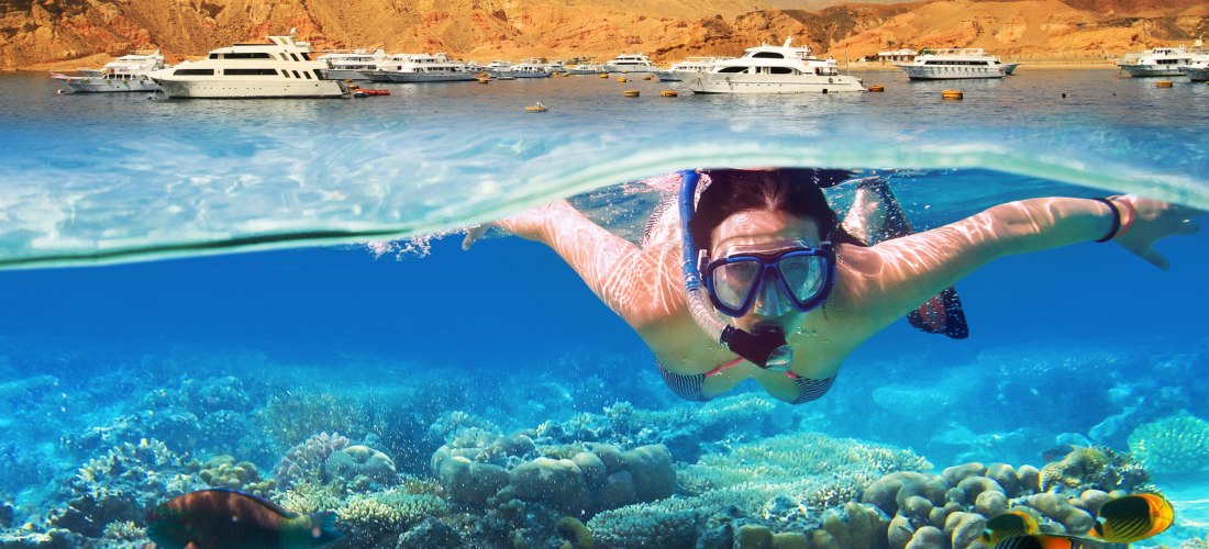 Frau mit Schnorchelmaske schwimmt unter Wasser zwischen bunten Korallen und tropischen Fischen. Im Hintergrund sind mehrere weiße Yachten vor einer felsigen Küste mit rotbraunen Bergen zu sehen.