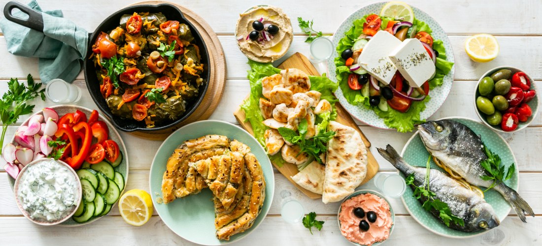 Bunte Auswahl an griechischen Vorspeisen und Gerichten auf einem weiß gedeckten Tisch: darunter gefüllte Weinblätter, gegrillter Fisch, Hummus, Tzatziki, griechischer Salat mit Feta, gegrilltes Hähnchen, Fladenbrot, gefüllte Paprika, Oliven und Ouzo.